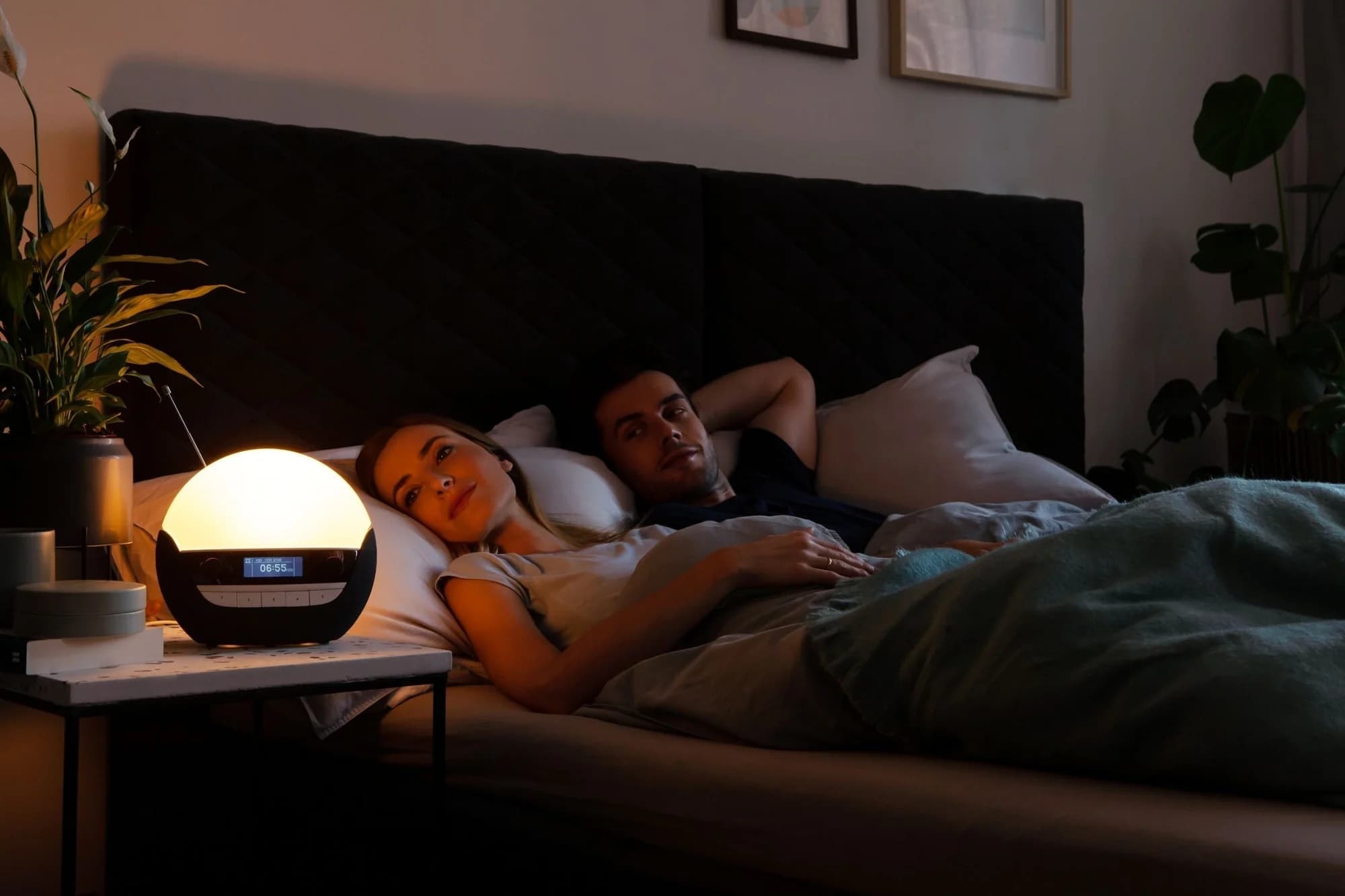 Der Lichtwecker Lumie 750 DAB Luxe leuchtet warm auf dem Nachttisch, daneben ein Paar im Bett
